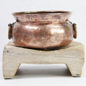 May include: A copper planter with two decorative handles sits on a wooden stand. The planter has a hammered finish and is slightly tarnished.