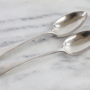 May include: Two silver-toned metal spoons with oval bowls and slender handles. Each handle features a small, engraved monogram at the rounded end. The spoons have a polished, reflective surface and a smooth, simple design throughout the entire length.