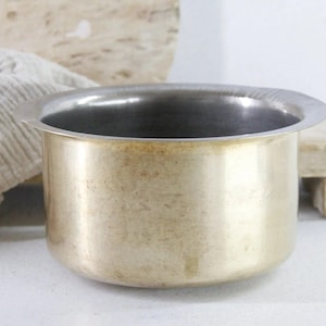 May include: A cylindrical brass pot with a wide, flat rim and a smooth, metallic finish. The interior of the vessel is silver-toned, contrasting with the warm, golden-yellow exterior surface of the metal container, which features a simple, unadorned design.
