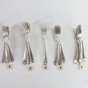 May include: A collection of silver-toned metal forks arranged in seven distinct groups, labeled A through G with small wooden tiles. Each group contains multiple forks with a classic design, featuring four tines and a smooth, curved handle.