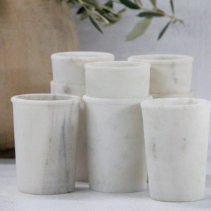 May include: A set of six white marble cups, stacked and arranged in a group. The cups are smooth and have a slightly textured surface.
