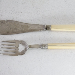 May include: A silver fish knife and fork set. The knife has a wide, ornate blade with a decorative pattern. Both pieces have cream-colored handles and silver accents. The set is displayed on a white surface.