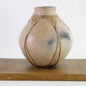May include: A beige ceramic vase with a rounded body and a narrow neck, adorned with vertical, light brown accents. The vase rests on a vintage book with a worn, tan cover. The overall aesthetic is rustic and handcrafted.