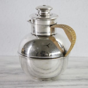 May include: A polished silver-toned metal pitcher with a spherical body, a short spout, and a removable lid featuring a small knob handle. The pitcher includes a curved handle wrapped in light tan woven rattan material, providing a textured contrast to the smooth, reflective metallic surface of the vessel.