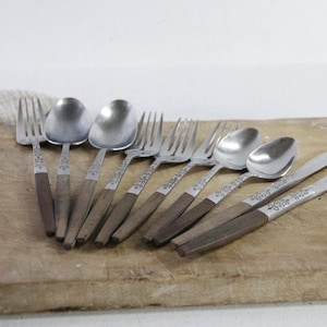 May include: A collection of vintage silverware, including forks, spoons, and knives, with wooden handles and decorative metal accents. The utensils are arranged on a wooden cutting board, showcasing a rustic aesthetic.