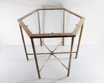 May include: A hexagonal side table featuring a clear glass top set within a metallic gold-toned frame. The table has four legs designed to resemble bamboo stalks, connected by a central X-shaped support brace at the base of the structure.