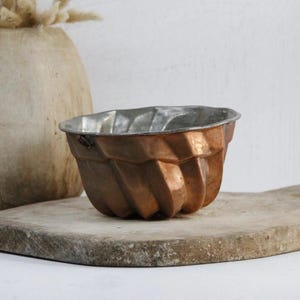 May include: A copper-colored, fluted, vintage cake mold sits on a rustic wooden cutting board. The mold is made of metal and has a slightly worn appearance.