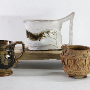 May include: A set of three ceramic mugs with a speckled, matte finish. The mugs are in shades of cream, light gray, and dark gray. Each mug has a simple, modern design and a rounded handle.