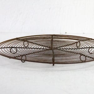 May include: Oval-shaped, antique metal cooling rack. The rack is made of thin, concentric wires with a handle on one end. The metal has a rustic, aged appearance, with a dark brown color. The design features circular accents.
