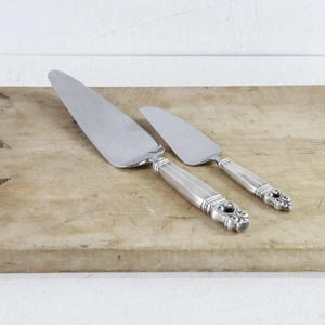 May include: A silver-toned cake server and a smaller matching knife with ornate, detailed handles. The utensils feature polished metal blades and are positioned diagonally on a rectangular, light-colored wooden cutting board with a handle.