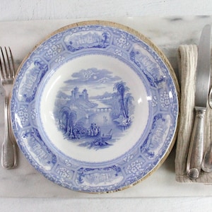 May include: A blue and white ceramic soup plate featuring a detailed transferware landscape scene with a castle, river, and figures. The plate is accompanied by a silver-toned fork, a silver-toned knife, and a silver-toned spoon on a textured napkin.