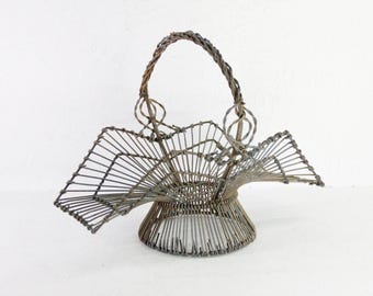 May include: A vintage metal wire basket with a handle. The basket has a flared base and a decorative fan-like design on the sides. The handle is made of twisted wire. The basket is a neutral gray color.