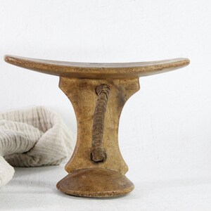 May include: A handcrafted wooden stool with a curved seat and a decorative handle. The stool is light brown with a textured surface and a rounded base. The handle is made of a twisted rope-like material.