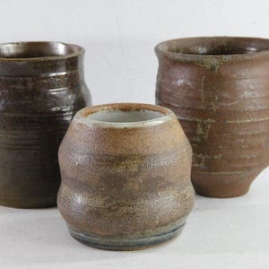 May include: Three handcrafted ceramic vessels in varying sizes and shapes. The pottery features earthy brown tones with textured surfaces. The largest vessel is cylindrical, the middle one is rounded, and the smallest is a squat shape.