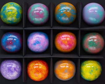 Op de afbeelding: Twaalf ronde, kleurrijke, beschilderde chocoladesferen gerangschikt in een rasterpatroon in een zwarte doos. De chocolaatjes hebben een verscheidenheid aan kleuren en patronen, waaronder geel, blauw, groen, paars en oranje.
