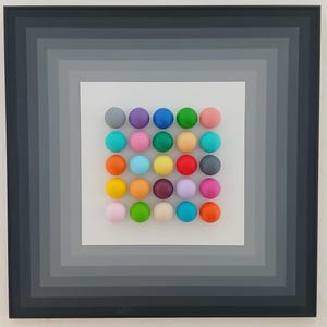May include: A framed artwork featuring a white background with a grid of 25 colorful spheres. The spheres are arranged in a 5x5 grid and are painted in various shades of red, orange, yellow, green, blue, purple, pink, and brown. The artwork is framed with a black frame with a series of concentric gray squares.