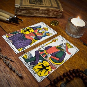 Puede incluir: Dos cartas de tarot etiquetadas como L'IMPERATRICE y L'EMPEREUR con ilustraciones coloridas de una reina y un rey. Las cartas se muestran junto a una vela encendida en un portavelas de cristal, libros antiguos, herramientas de tallado, lavanda seca y un rosario.