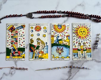 May include: Four tarot cards with colorful illustrations and French titles are displayed with a rosary and dried lavender sprigs. The cards feature images of a tower, a woman, a moon, and a sun with figures. The rosary is made of dark brown beads.