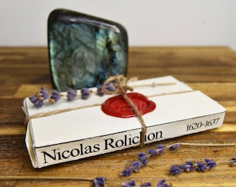 May include: A white book with a red wax seal and twine tied around it. The book is titled "Nicolas Rolichon 1620-1637" and is sitting on a wooden surface with dried lavender flowers.
