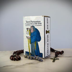 Puede incluir: Una caja de cartas del tarot con el texto "Tarot Piemontais Cesare Riva 1841-1846" y una ilustración de una figura vestida con una túnica sosteniendo una linterna. Un rosario y una ramita de lavanda están en primer plano. La caja es blanca con detalles florales azules.