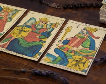 May include: Three tarot cards on a wooden table. The cards are from a vintage deck and feature colorful illustrations of figures and symbols. The cards are labeled "Imperator", "Gem Perever", and "4."