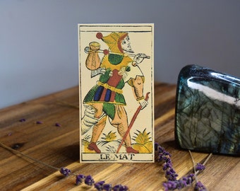 May include: A tarot card with a colorful illustration of a jester holding a staff and a bag. The card is labeled "Le Mat" and is on a wooden surface with lavender flowers and a blue-green crystal.