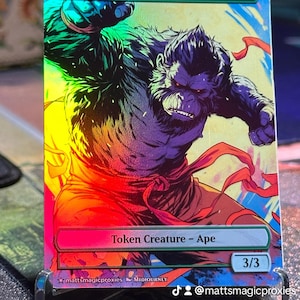 May include: A holographic Magic: The Gathering card featuring an illustration of an ape with a red sash. The card text reads "Ape" and "Token Creature - Ape" with a power and toughness of 3/3.