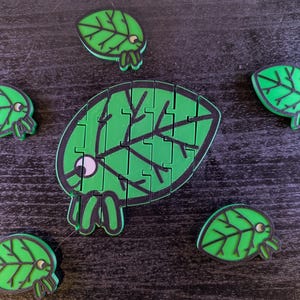 Puede incluir: Cinco piezas de rompecabezas en forma de hoja verde con contornos negros y ojos de dibujos animados. Las piezas están dispuestas sobre una superficie oscura. La pieza más grande es un rompecabezas con múltiples secciones.