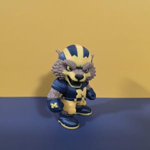 May include: A small figurine of a wolf-like character wearing a dark blue and yellow football uniform. The character has a yellow and blue helmet, a dark blue jersey with a yellow letter M on the chest, dark blue gloves with a yellow M, and boots.