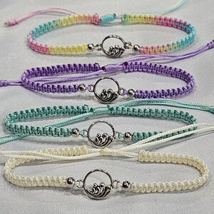 May include: Macrame friendship bracelets in pastel colors: rainbow, purple, teal, and cream. Each bracelet features a silver-toned wave charm and adjustable sliding knot closure. These handmade bracelets are perfect for everyday wear.