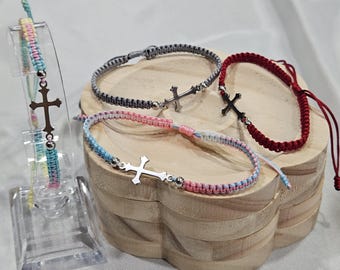 May include: Four adjustable bracelets featuring silver-toned cross charms and metallic beads. One bracelet is displayed on a clear stand, while three others are arranged in a group. The cords are woven in various colors including red, grey, and pastel blue, pink, and yellow combinations.