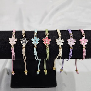 May include: A collection of seven flower charm bracelets in various colors, including pink, black, blue, green, white, purple, and another pink. Each bracelet features a braided cord and gold-colored accents. The flower charms are the focal point.