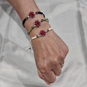 May include: Three adjustable bracelets with red rose charms. The bracelets are black, green, and off-white, with silver-colored beads. The rose charms are a vibrant red with silver-colored metal detailing. The bracelets are displayed on a wrist.