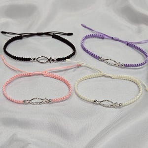 May include: Four macrame bracelets in black, purple, pink, and white. Each bracelet features a silver fish charm and adjustable sliding knots. The bracelets are displayed on a white fabric surface.