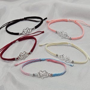 May include: Five handmade friendship bracelets in various colors: black, pink, red, cream, and a blue/pink mix. Each bracelet features a silver heart-shaped charm and adjustable macrame knot closure.