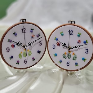 May include: Two white embroidery hoop clocks with black hands and Roman numeral markings. The clocks are decorated with colorful embroidered flowers and are displayed on clear plastic stands.