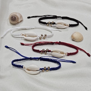 May include: Five macrame bracelets in black, white, red, and blue, each featuring a cowrie shell and gold-colored beads. Two seashells are placed near the bracelets. The bracelets are on a light green surface.
