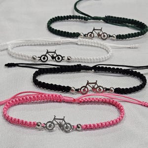 May include: Four adjustable macrame bracelets featuring a central silver-toned bicycle charm. Each bracelet is crafted from cord in distinct colors: dark green, white, black, and pink. Small metallic beads flank the bicycle charm on each cord.