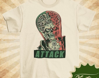 May include: Cream-colored t-shirt featuring a vintage graphic of a Martian alien with a skull-like face and exposed brain. The word "ATTACK" is printed in green block letters below the alien. The shirt has short sleeves.