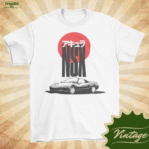Könnte beinhalten: Weißes T-Shirt mit einer Grafik eines silbernen Sportwagens und dem schwarzen Text "NSX" vor einem roten Kreis, mit japanischen Schriftzeichen darüber. Das Design hat einen Vintage-Stil.