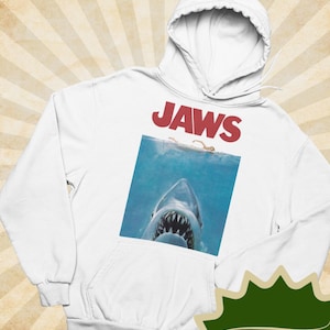 May include: A white hooded sweatshirt featuring a graphic print of the Jaws movie poster. The design includes the red text JAWS above an illustration of a shark swimming towards a person floating on the surface of the blue ocean water.