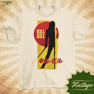 May include: A cream-coloured t-shirt with a graphic from the film "Kill Bill." The design features a black silhouette of a person with a sword, set against a red circle and yellow background. The words "KILL BILL" and Japanese text are also present.