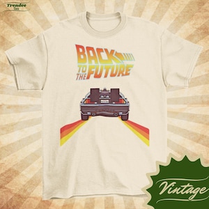 Könnte beinhalten: Ein weißes T-Shirt mit einem Vintage-Grafikdruck des DeLorean-Zeitfahrzeugs aus dem Film "Zurück in die Zukunft". Die Grafik zeigt den Text "Zurück in die Zukunft" und ein stilisiertes Bild des DeLorean, der auf einer Straße mit gelben Streifen fährt.