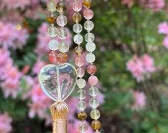 May include: A beaded necklace with a clear heart-shaped pendant and a tassel. The necklace features a variety of colorful beads in shades of pink, yellow, and brown. The tassel is a light brown color. The background is a blurred view of pink flowers.
