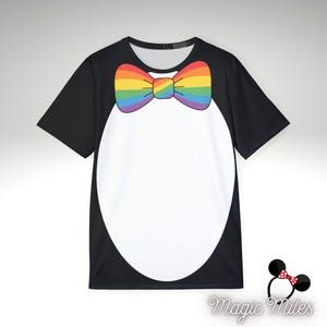 Pode incluir: Uma t-shirt preta de manga curta com um grande oval branco na frente e um gráfico de laço com riscas arco-íris no decote. A t-shirt tem um decote redondo e um acabamento liso. Um pequeno logótipo com orelhas de rato e o texto Magic Miles.