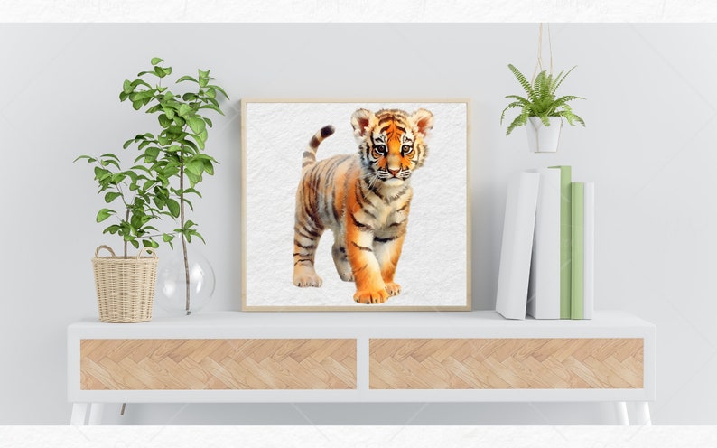 K&ouml;nnte beinhalten: Ein Aquarellbild eines niedlichen Tigerbabys mit orangefarbenen und schwarzen Streifen, das auf einem wei&szlig;en Hintergrund steht. Das Bild ist in einem hellen Holzrahmen gerahmt und auf einem wei&szlig;en Regal mit einer Holzplatte ausgestellt.