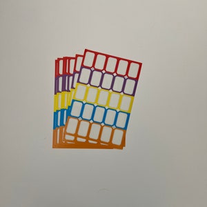 May include: A stack of four sheets of colorful sticker labels. Each sheet has a grid of 20 white rectangular labels with a black border. The sheets are red, purple, yellow, and blue.