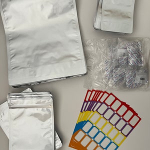 May include: A stack of silver mylar bags with a zipper closure, a stack of smaller silver mylar bags with a zipper closure, a clear plastic bag filled with small white silica gel packets, and a stack of colorful rectangular labels with white blank spaces.