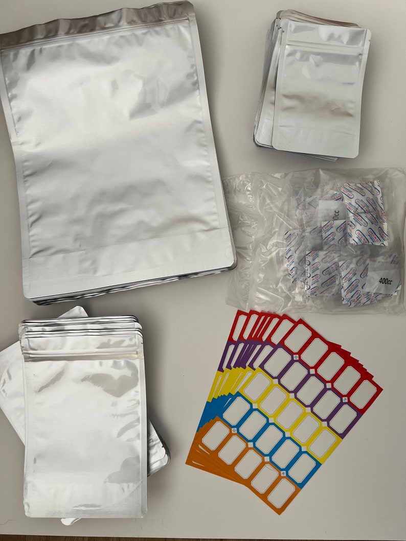 May include: A stack of silver mylar bags with a zipper closure, a stack of smaller silver mylar bags with a zipper closure, a clear plastic bag filled with small white silica gel packets, and a stack of colorful rectangular labels with white blank spaces.