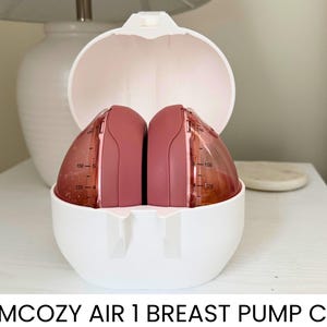 May include: A white breast pump case with a hinged lid, open to reveal two pink breast pump components. The components have measurement markings. The text "MOMCOZY AIR 1 BREAST PUMP CASE" is at the bottom of the image.